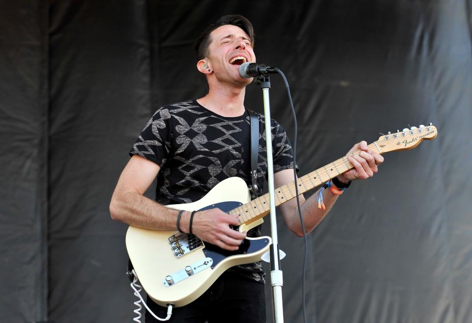 A man wearing a black patterned crewneck shirt sings and plays electric guitar.