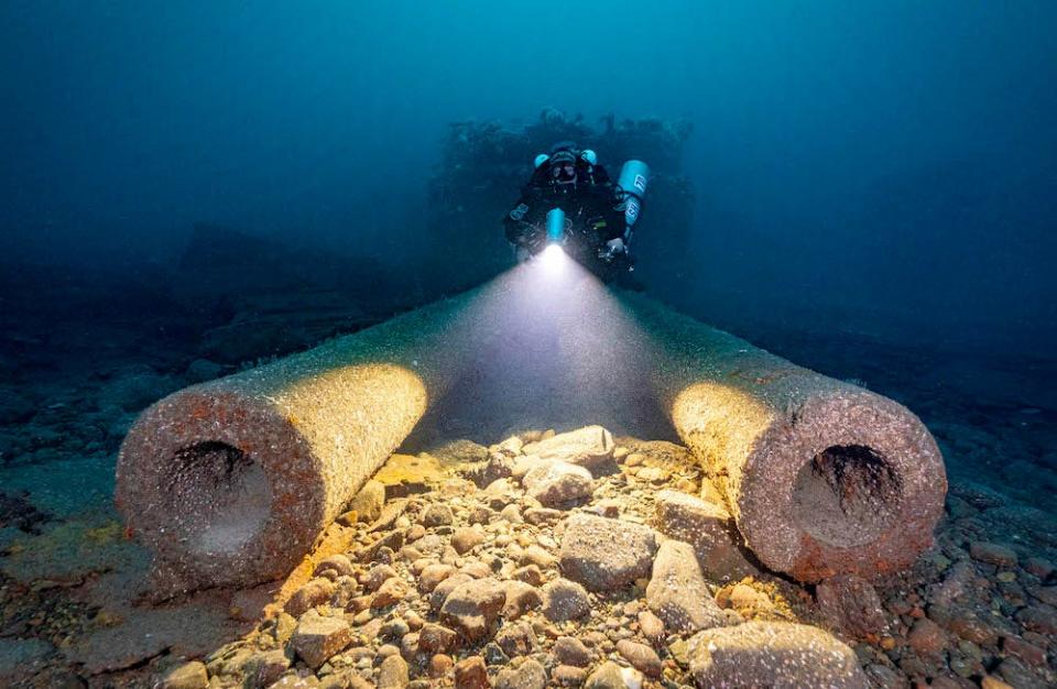 A diver faces the camera with two gun barrels of the HMS Audacious, sunk off the coast of Ireland, in the foreground. The image is a winner in the 2024 Underwater Photographer of the Year Awards.