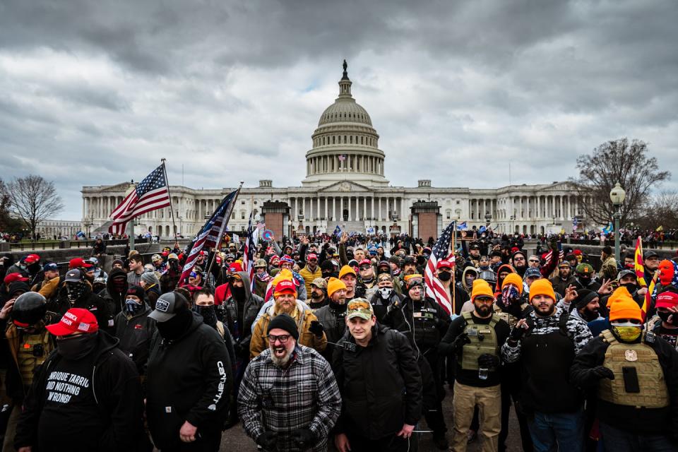 A crowd of angry people in front of a large white, domed building, with dark clouds above.