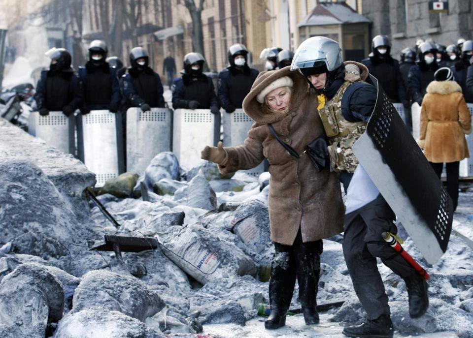A protester wearing improvised protective gear helps a woman cross near the barricade in front of riot police in Kiev, Ukraine, Friday, Jan. 24, 2014. Protesters have seized a government building in the Ukrainian capital while also maintaining the siege of several governors' offices in the country's west, raising the pressure on the government. After meeting with President Viktor Yanukovych on Thursday, opposition leaders told the crowds that he has promised to ensure the release of dozens of protesters detained after clashes with police and stop further detentions. (AP Photo/Darko Vojinovic)
