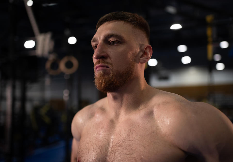 A muscular man with short hair and a beard stands shirtless in a gym, looking determined