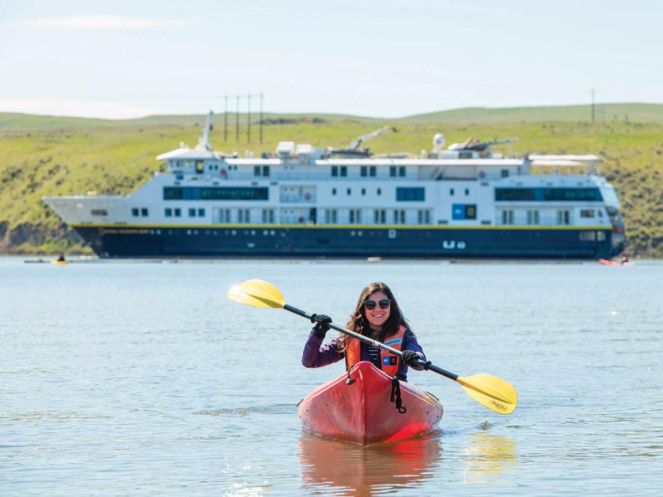 A person kayaking away from a large cruise ship on a green coast.