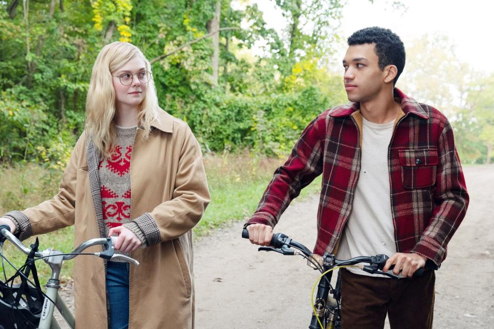 Elle Fanning stands outside, walking her bicycle; she looks at a boy walking next to her, also walking his bicycle