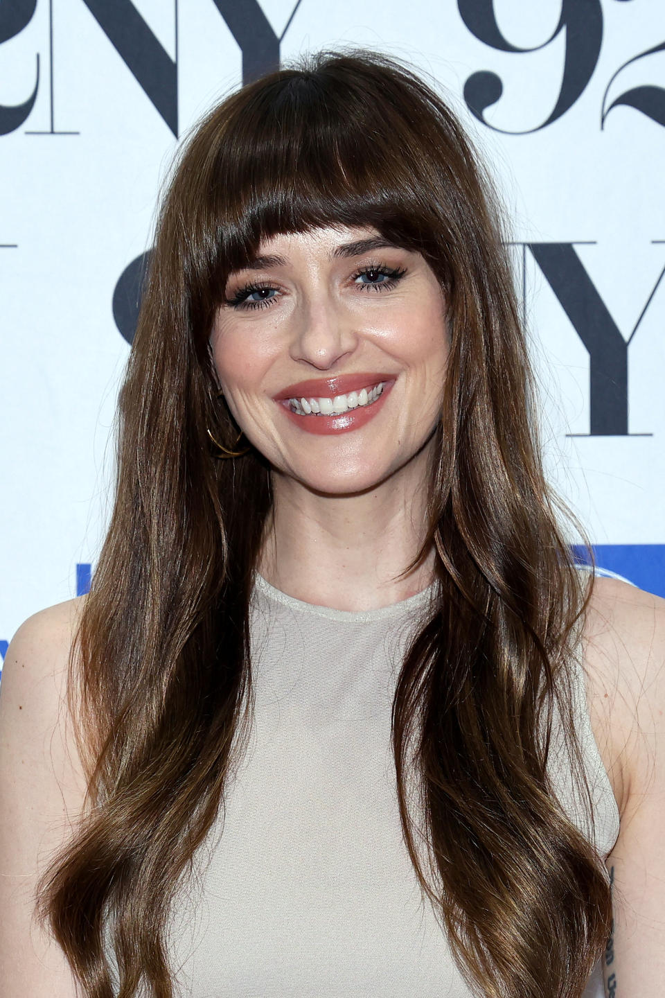 Dakota Johnson smiling in front of a backdrop with partial text and logos. She is wearing a sleeveless, high-neck top