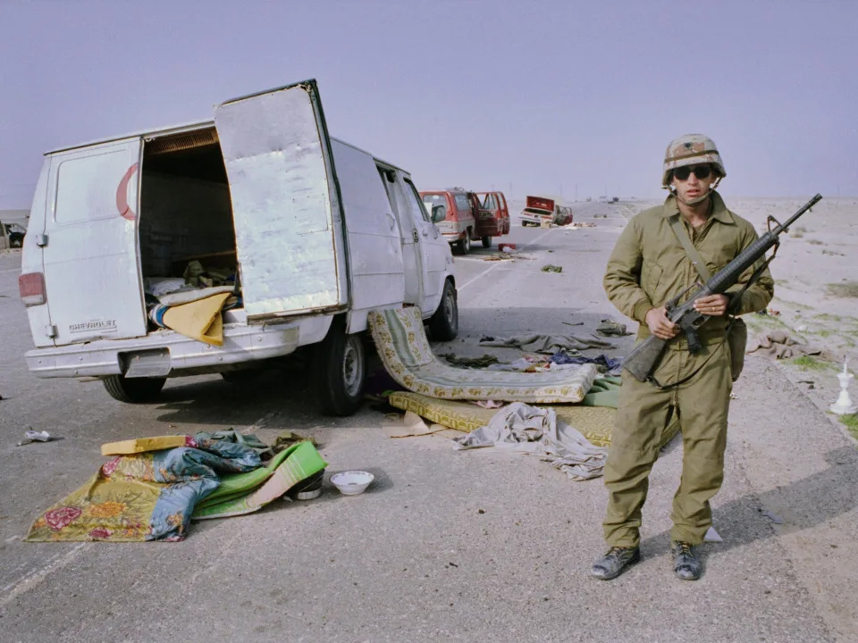 A US soldier holding an M16 rifle beside abandoned vehicles with their doors open in Saudi Arabia in 1991.