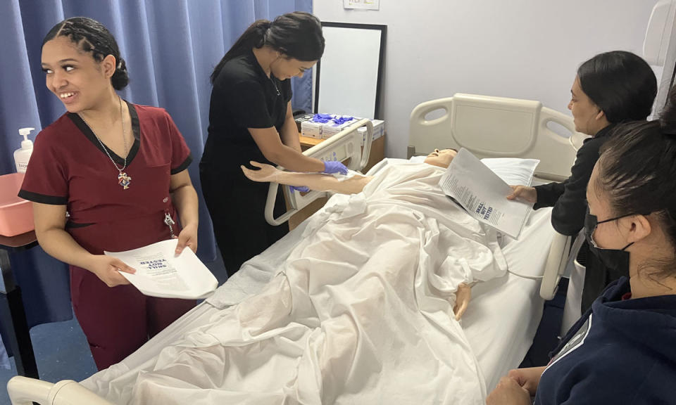 Diana Pimentel (left) listens to an advisor as RINI classmates (from left) Veronica Benitez, Joslin Lebron and Edilma Ramirez tend to a mock patient in a prep session for a certified nursing assistant exam. (Greg Toppo)