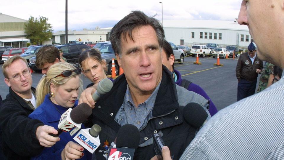 mitt romney wearing a jacket, talking to reporters holding several microphones in front of him as they stand in a parking lot outside