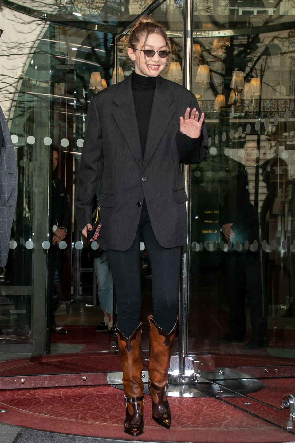 <p>In an oversized black blazer, black turtleneck, black jeans, angular sunglasses, and brown and black leather boots. </p>