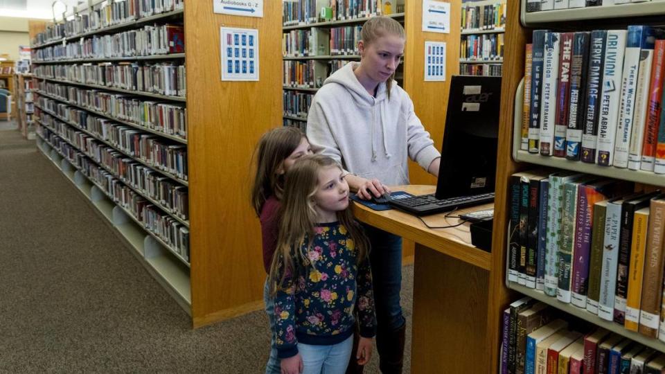 Andrea Kitzmiller, 5, and older sister Claire, 7, watch as their mother, Rachel Kitzmiller, uses a computer terminal to search for a book at the Meridian Library in March 2023. Some libraries are restricting children’s access to materials through distinct library cards, “adults only” sections or parental consent.