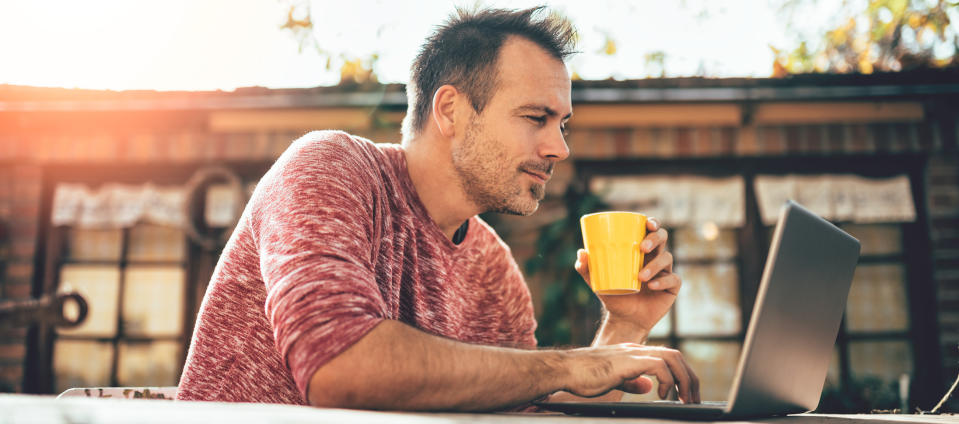 Man sitting in front of his laptop outdoors holding a drink in a yellow cup.