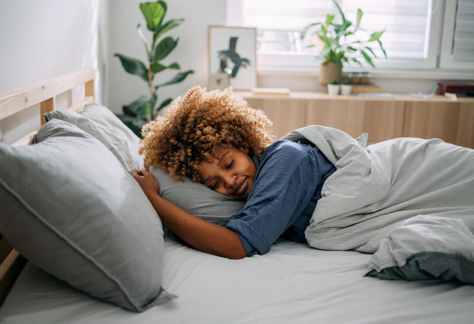 A curly-haired man sleeps comfortably in a bed with grey sheets in a bright bedroom with plants and framed paintings in the background.