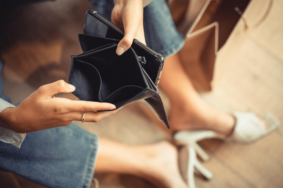 A person in jeans and sandals checks an empty black wallet. The photo emphasizes financial uncertainty or budgeting