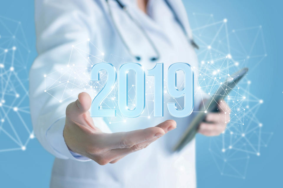 Physician with stethoscope around neck holding tablet in one hand and extending other hand with an image of 2019 appearing above the hand and connected points of light in the foreground