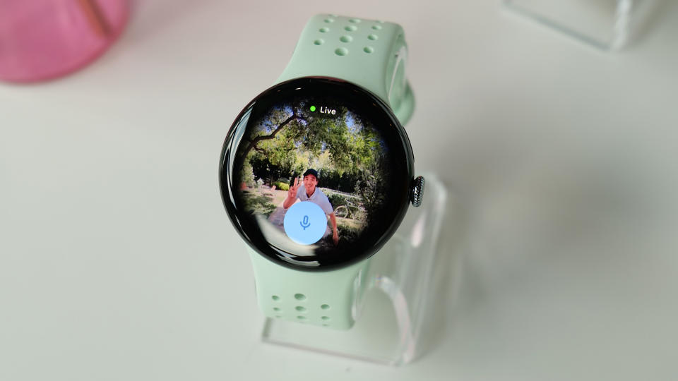 A Pixel Watch 3 on a stand with a video feed on its screen. A person appears to be smiling and waving at a camera, and a microphone is overlaid on top of the image in the watch.