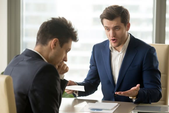 Man in suit looking critically at a second professional male who's looking down.