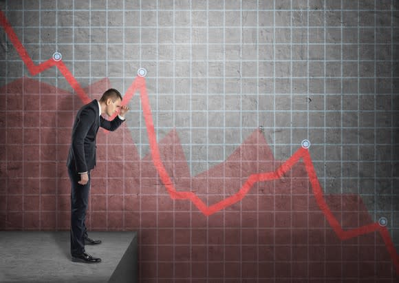 Man in a suit staring down into a drop off with a red, downward trending chart in the background.