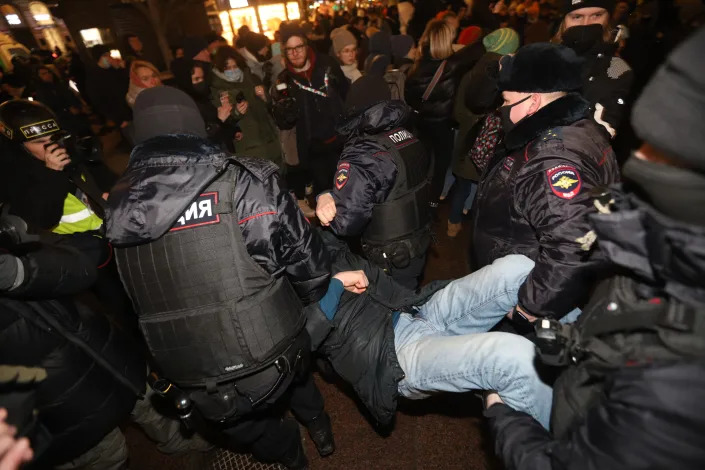 Police officers in black masks and black fur hats bodily remove a protester as demonstrators look on.