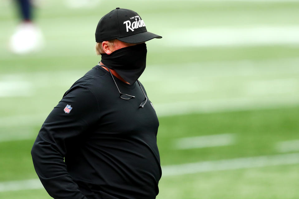 Jon Gruden stands on the field before the game against the Patriots with a full mask covering his face.