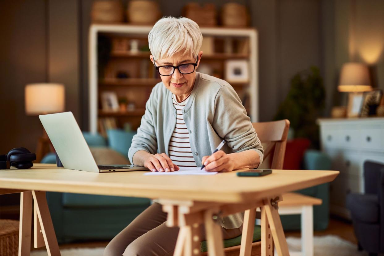 A diligent and focused senior adult female accountant with reading glasses working from home and writing financial reports in front of a laptop.