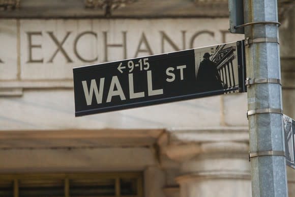 Wall Street road sign with a stock exchange sign on a stone building in the background.