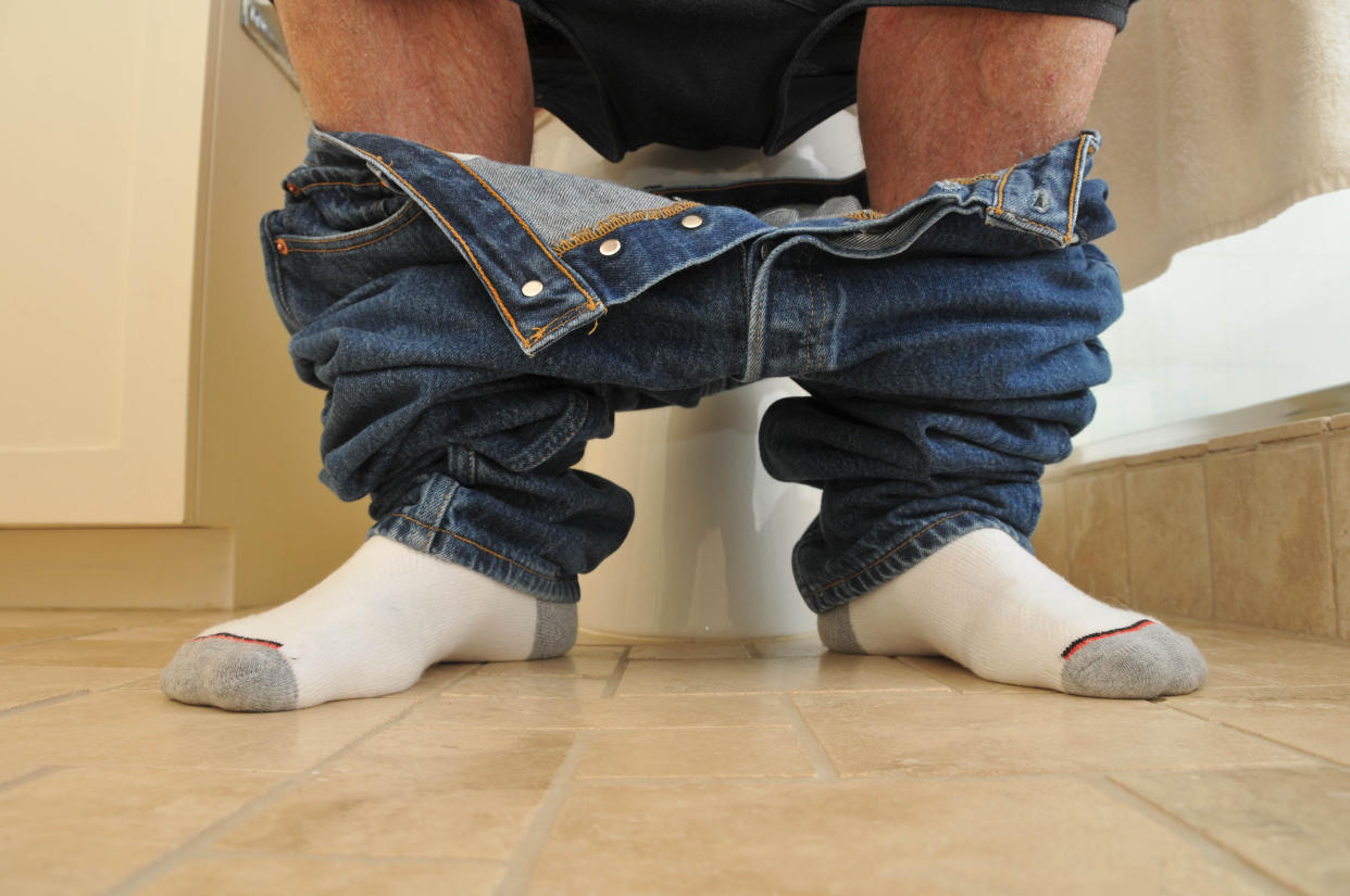 Jeans and underwear pushed down around a man’s calves and ankles as he sits on the toilet.