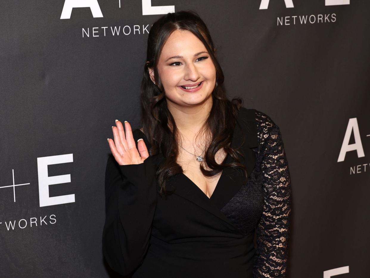 Gypsy Rose Blanchard in a black dress with lace sleeves smiles and waves to the camera in front of a black background.