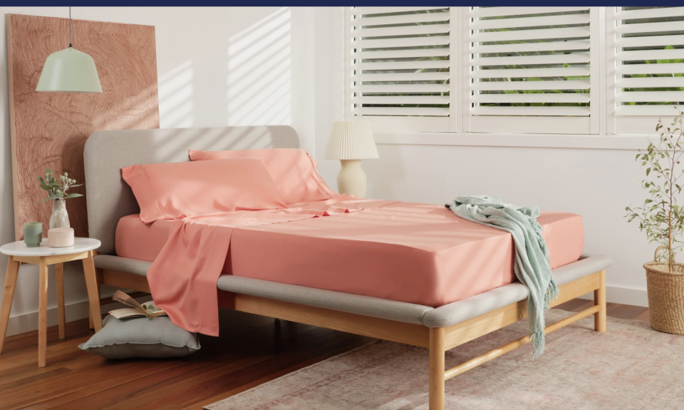 A bedroom scene showing coral coloured sheets and pillows on a wooden bed in a room with white louvered windows.