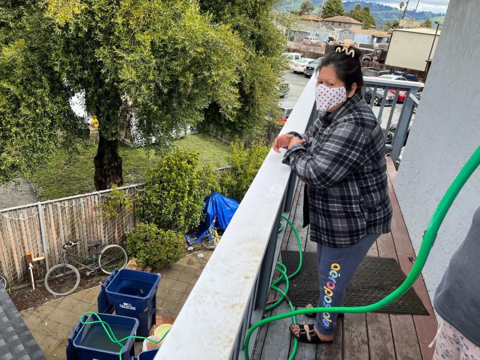 A woman stands on a balcony and watches two large blue trash cans below