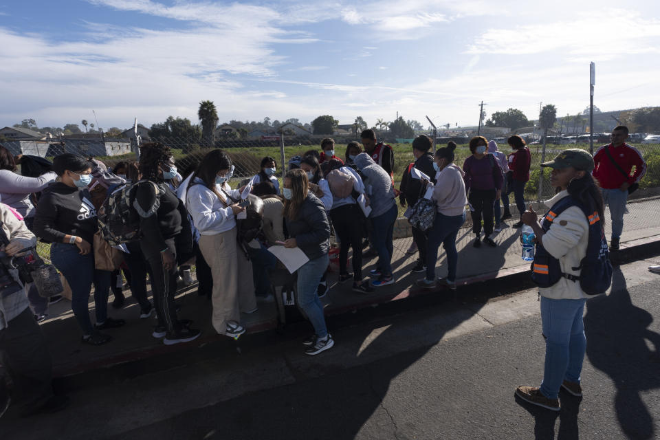 Migrants stand in groups as they arrive at a bus stop after leaving a processing facility, Friday, Feb. 23, 2024, in San Diego. Hundreds of migrants were dropped off Friday at a sidewalk bus stop amid office parks in San Diego with notices to appear in immigration court after local government funding for a reception center ran out of money sooner than expected. (AP Photo/Gregory Bull)