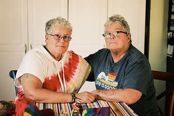 Gail Foreman and her wife, Pat Cummins, sitting at their kitchen table together. Cummins has her arm around Foreman. They both have short gray hair. Foreman has black wire-rim glasses and Cummins has rectangular dark frames.