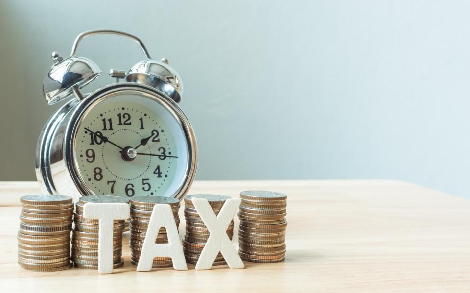 Piles of coins, alarm clock, and letter magnets spelilng TAX on a wood table in front of a light blue wall.