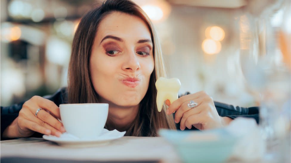 Mujer con una taza de café en una mano y una reproducción de un diente en otra.