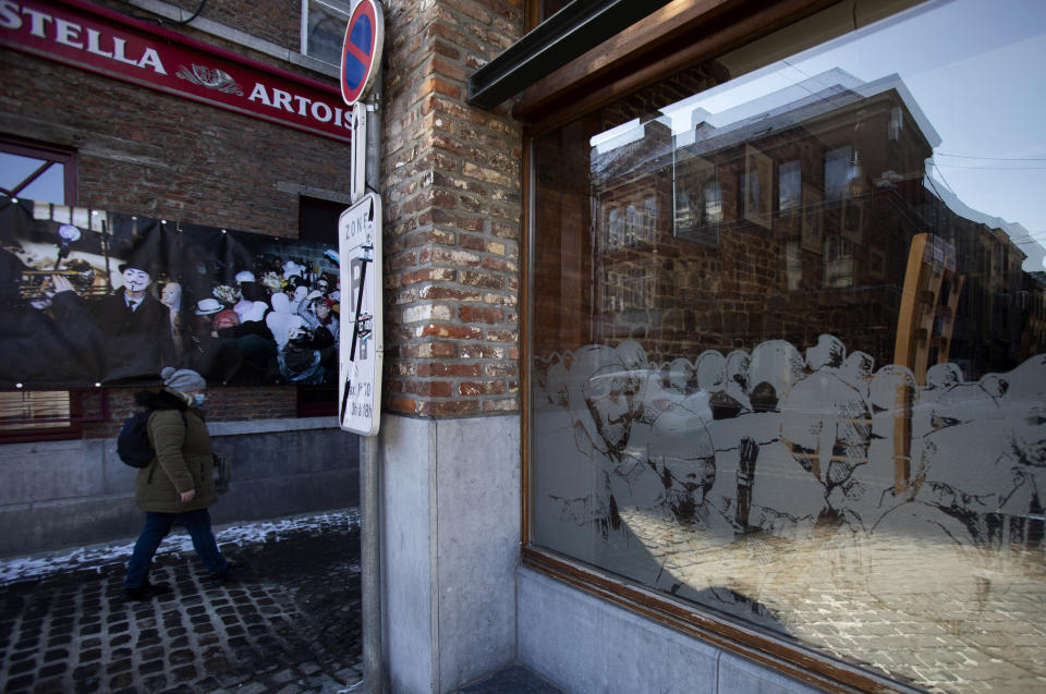 A woman walks past a carnival poster and a closed cafe that has chairs stacked in its window in the Grand Place of Binche, Belgium, Thursday, Feb. 11, 2021. In normal life, the small town of Binche in southern Belgium would be bursting with excitement for the carnival festivities that have been labeled a 'Masterpiece of the Oral and Intangible Heritage of Humanity' by UNESCO, but in 2021 the COVID-19 regulations have forced the carnival to be cancelled. (AP Photo/Virginia Mayo)