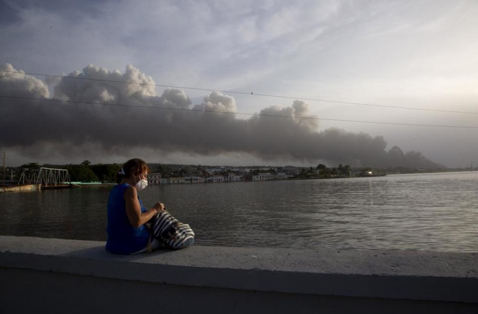 A resident sits on the malecon sea wall as smoke rises in the background from the fire.