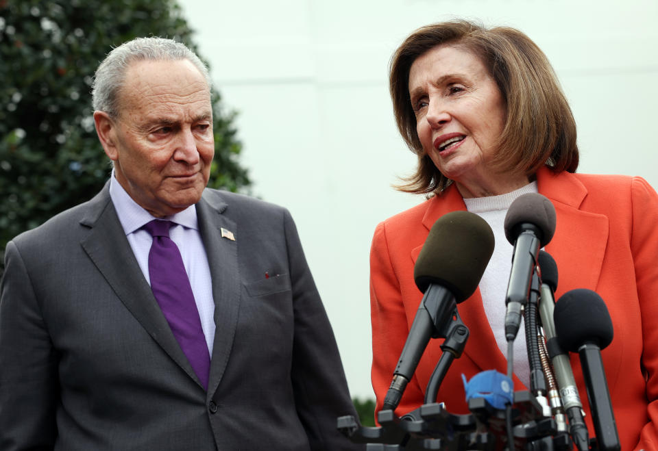 Chuck Schumer, in lavender shirt and purple tie, listens as Nancy Pelosi, in orange suit, speaks in front of a bank of microphones.