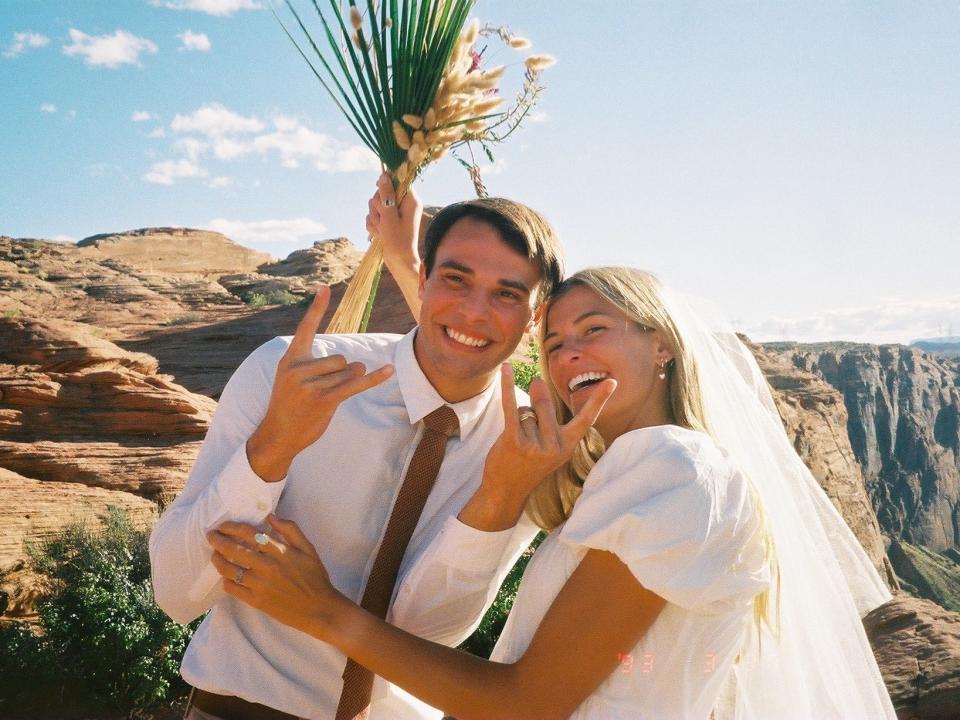 A couple. The woman is smiling and holding flowers, and the man is making a rockstar symbol with his hands.