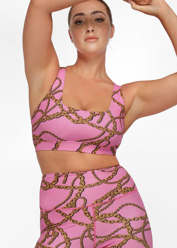 A model wears a pink sports bra and tights with gold chain motif. Linked Up Sports Bra