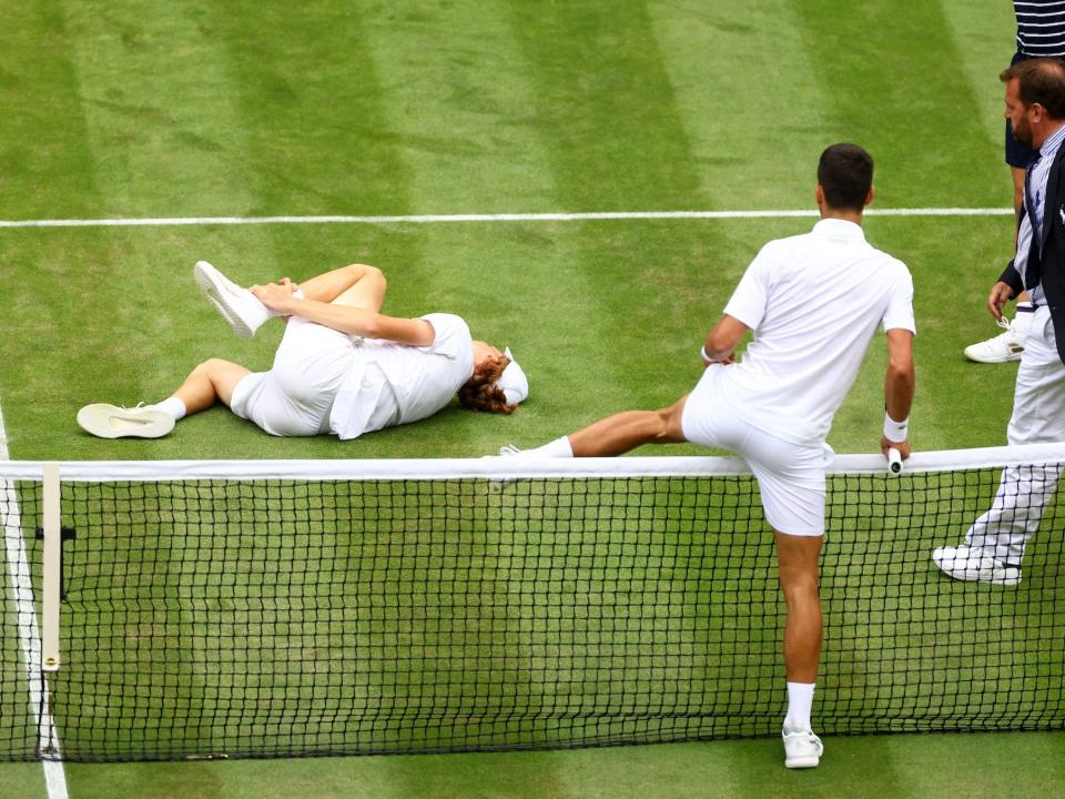 Novak Djokovic climbs over the net at Wimbledon while Jannik Sinner lays on the ground holding his ankle.