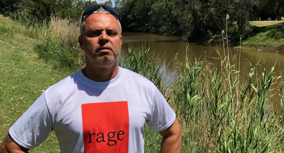 Steven Booby stands in front of the Namoi River. He wears a T-shirt that reads Rage.