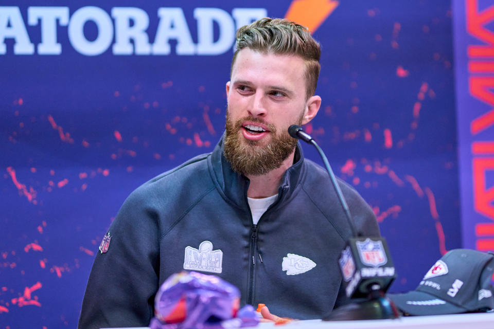 Harrison Butker with a beard, wearing a zip-up sports jacket, speaks into a microphone at a press conference with an NFL logo banner in the background