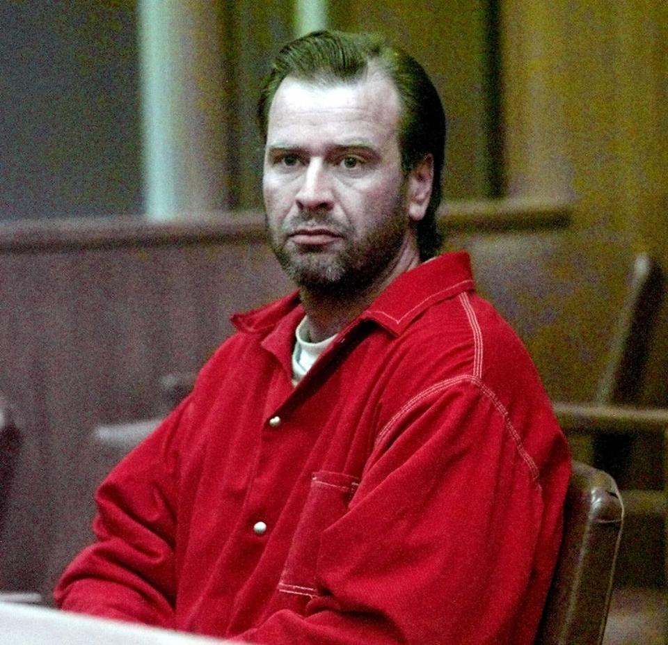 A square frame of a man with facial hair and wearing a red shirt, seated behind a table, looking slightly to the left