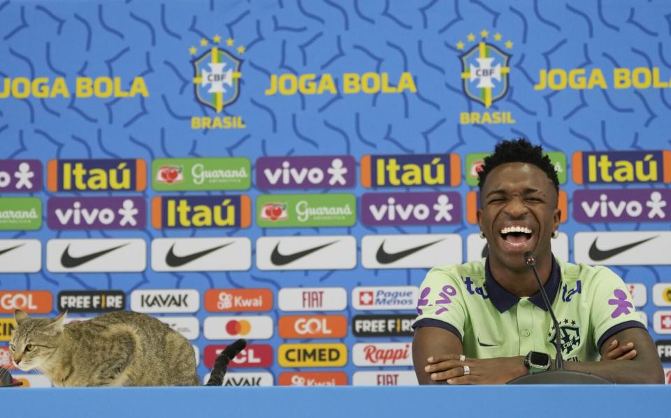 Vinicius Junior de Brasil se ríe mientras un gato se sienta en la mesa durante una conferencia de prensa.