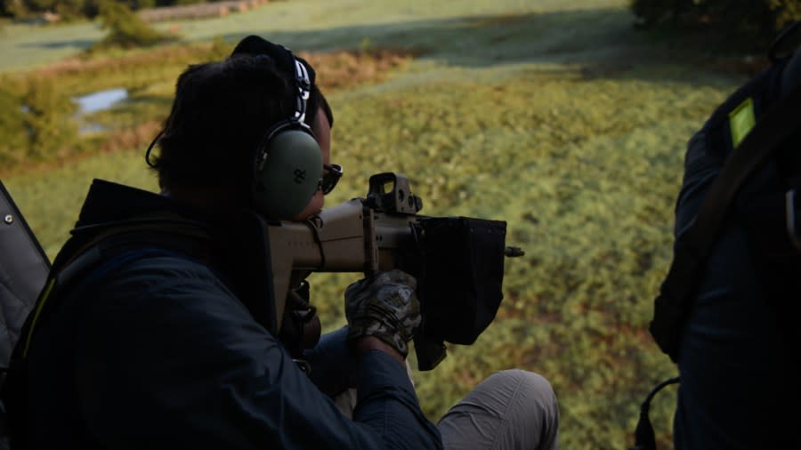 A man sits on the edge of a helicopter holding a large gun and earmuffs as he looks for feral hogs to shoot from the air.