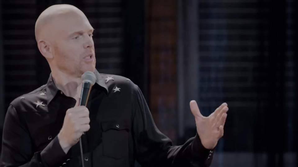 Bill Burr holding a microphone and holding one arm out on stage.