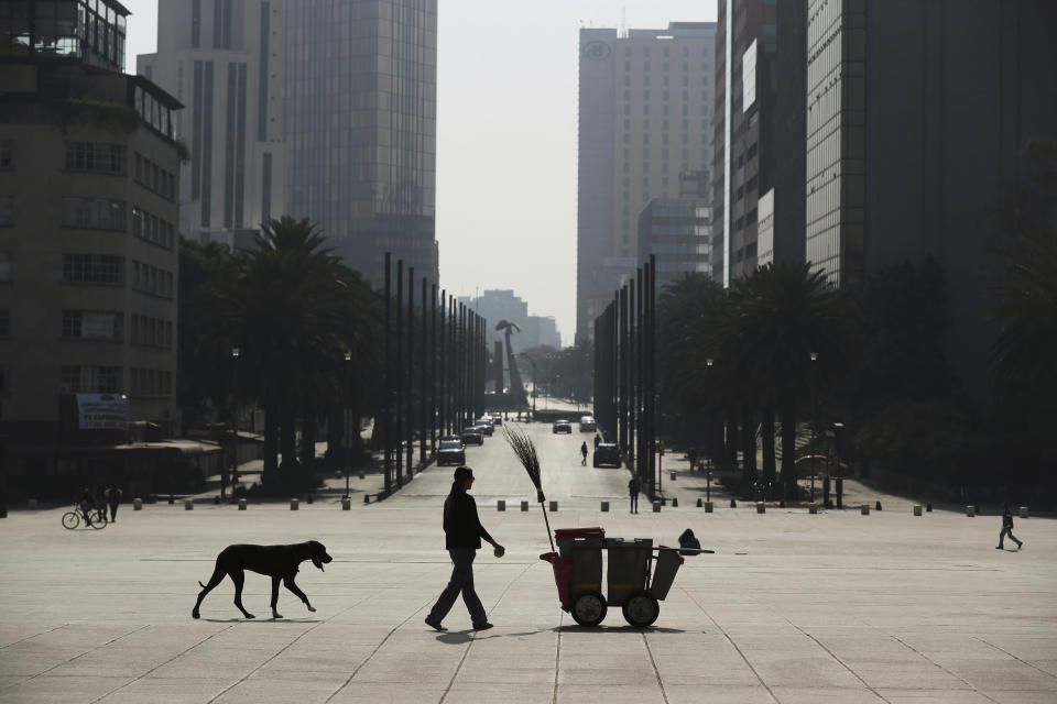 A woman walks her dog past a trash worker's cart, amid an empty plaza at Revolution Monument in Mexico City, Wednesday, April 1, 2020. Mexico's government has broadened its shutdown of "non essential activities," and prohibited gatherings of more than 50 people as a way to help slow down the spread of COVID-19. The one-month emergency measures will be in effect from March 30 to April 30. (AP Photo/Fernando Llano)