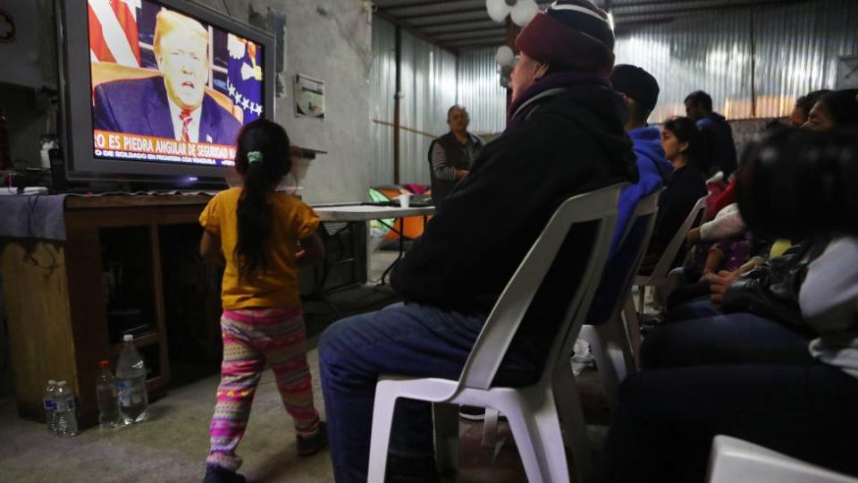 Un grupo de migrantes ven una transmisión televisiva donde aparece Trump en 2019.