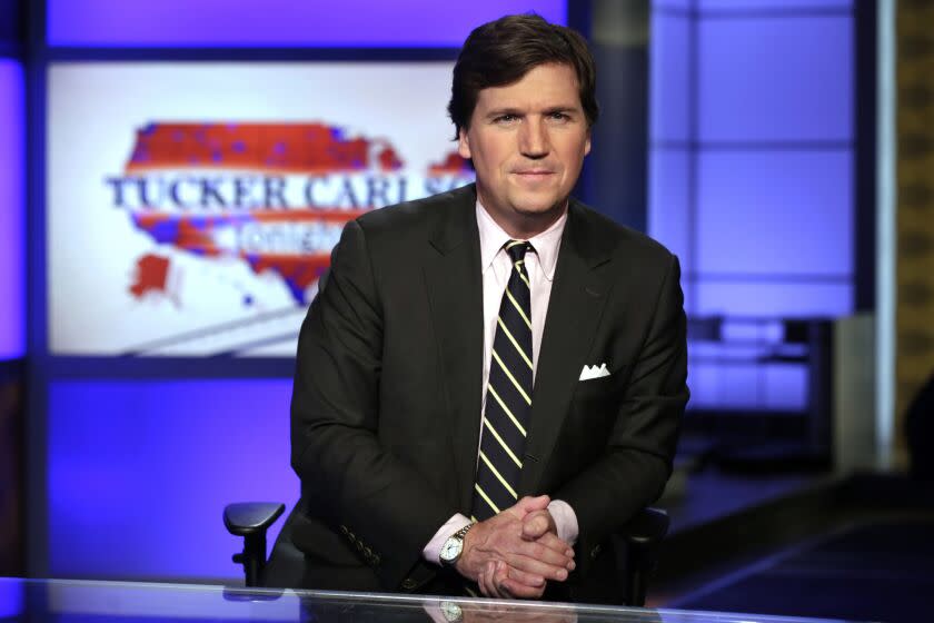 Tucker Carlson wears a suit and folds his hands on a desk