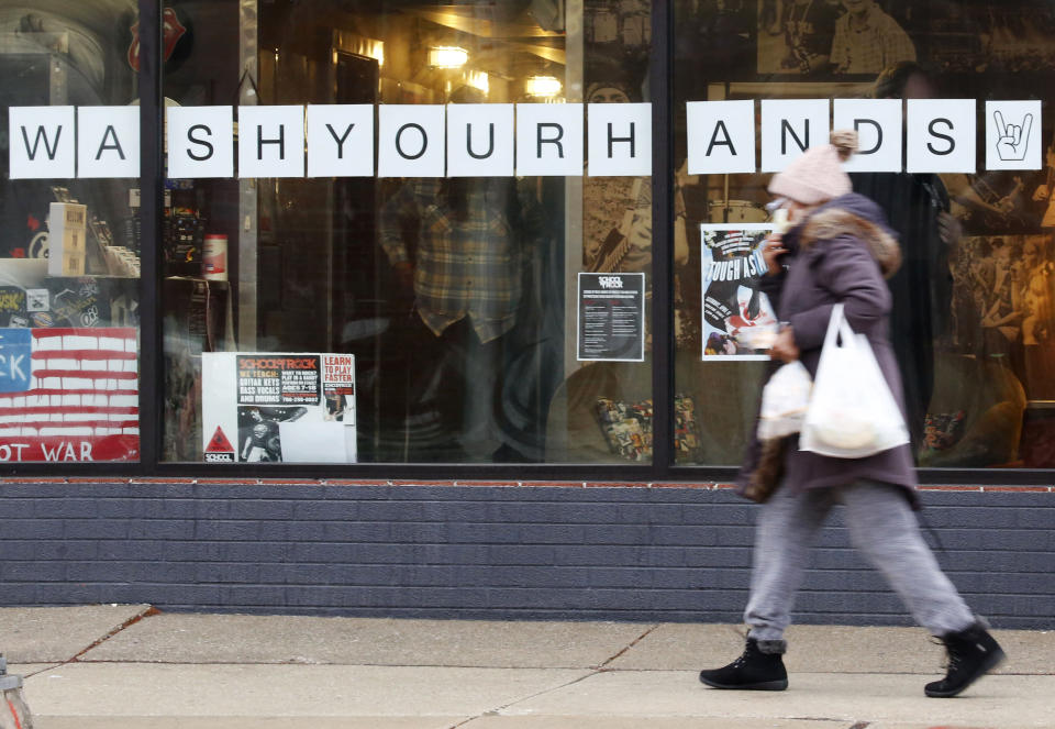 In this March 19, 2020, photo, a woman in a face mask walks past a "Wash Your Hands" sign after buying groceries in Oak Park, Ill., before a two-week "shelter in place" order goes into effect. Leaders in the Chicago suburb were the first in the state to take the step, requiring non-essential businesses to close, to try to curb the spread of the coronavirus. (AP Photo/Martha Irvine)