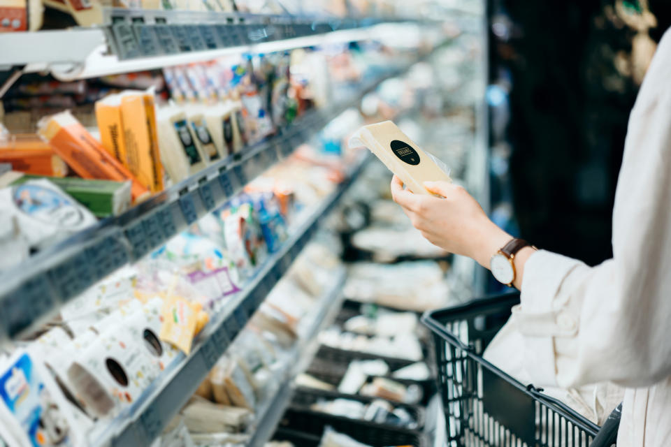 Cropped shot of young Asian woman with a shopping cart, shopping in the dairy section of a supermarket. She is reading the nutrition label on the package of a fresh organic cheese