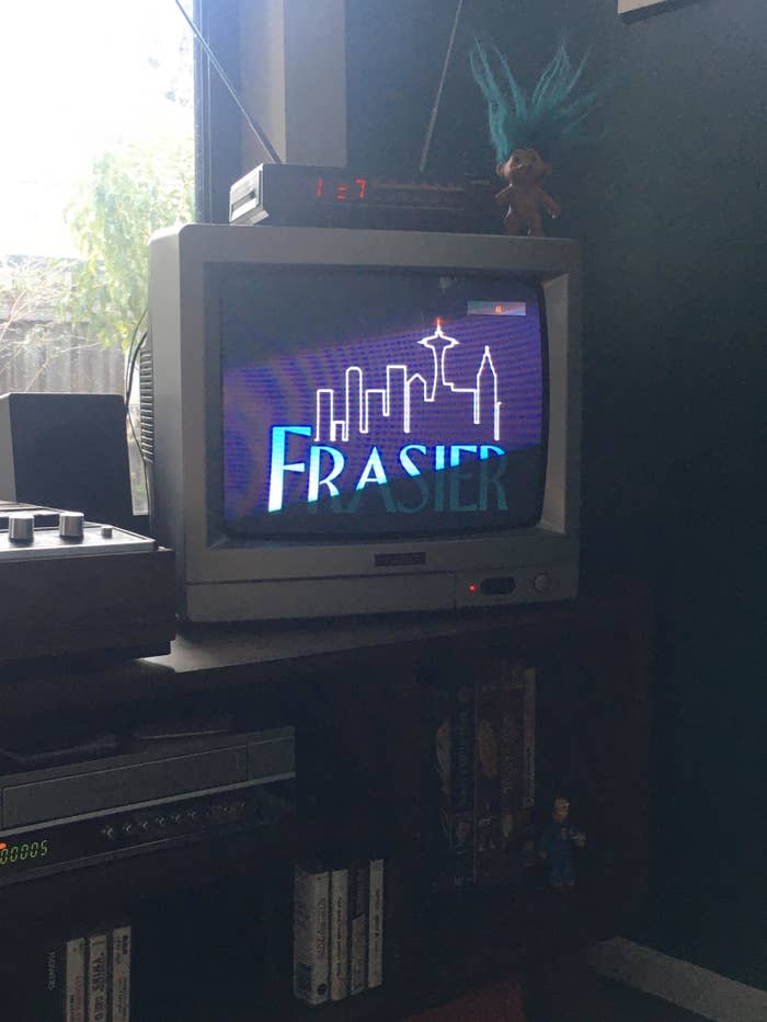 Old CRT TV displaying "Frasier" title screen, on a cabinet with gaming console, dolls nearby